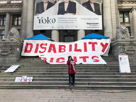 Me speaking into a mic infront of the steps at the BC museum. Behind me a large sign saying Disability Rights